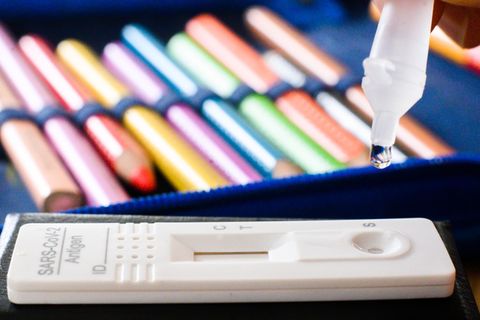 Ein klarer Tropfen hängt an einem Plastikröhrchen über einem Corona-Selbsttest. Im Hintergrund eine Federmappe mit Buntstiften