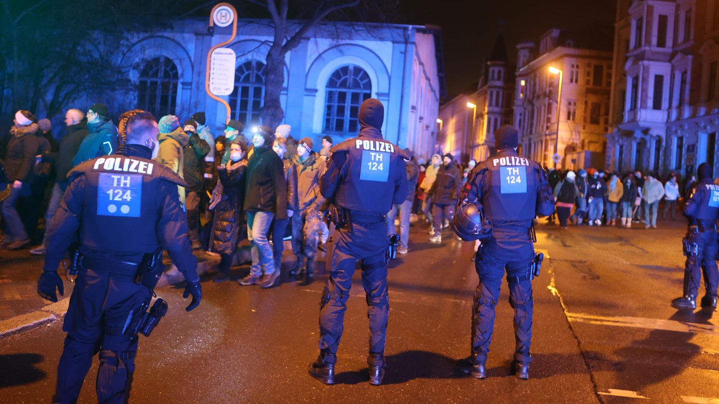 Drei Polizisten in dunkelblauen Uniformen sind von hinten zu sehen, während sie einen Demo-Zug beobachten