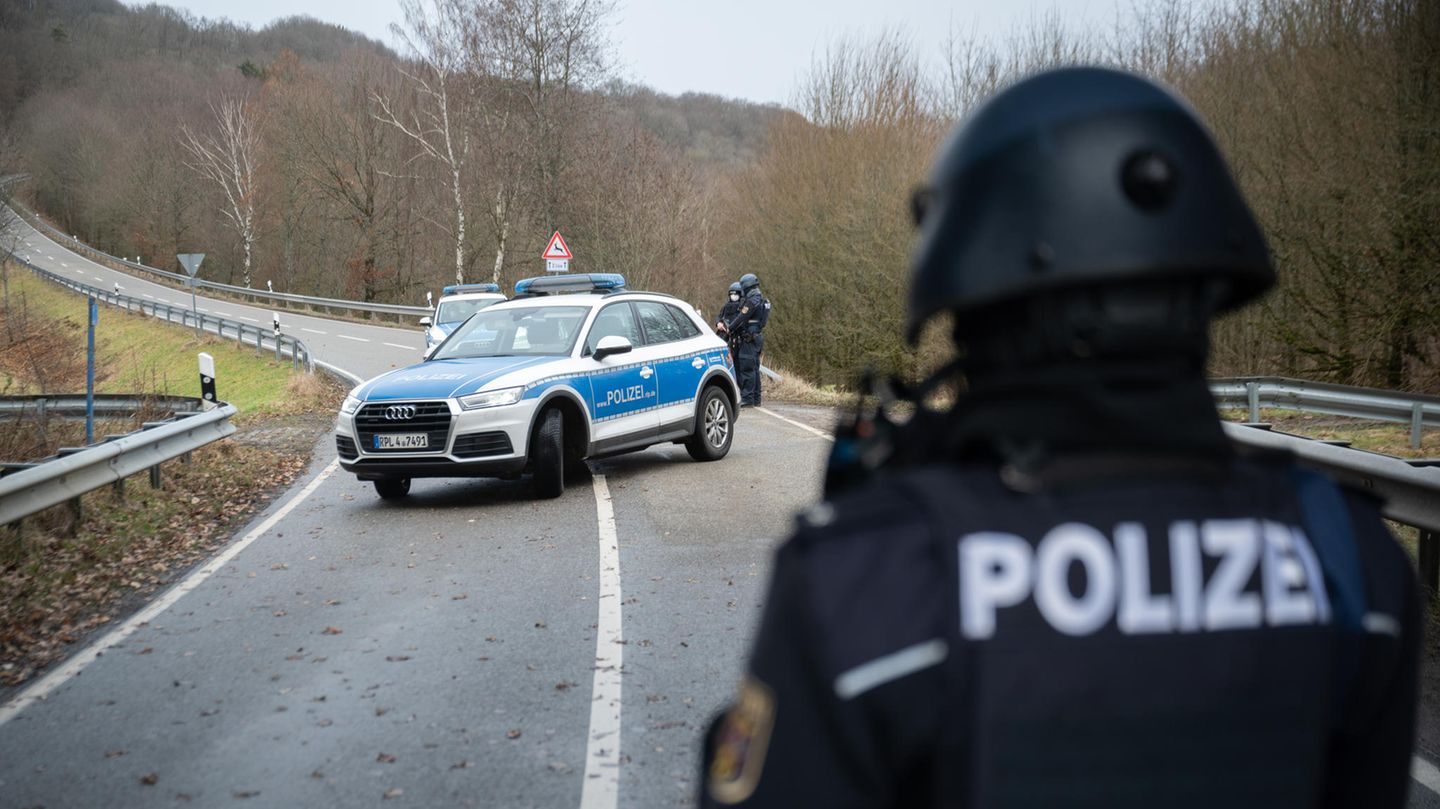 Ein Polizist in Schutzkleidung und mit Helmschaut auf ein diagonal über eine geschwungene Landstraße stehendes Polizeiauto