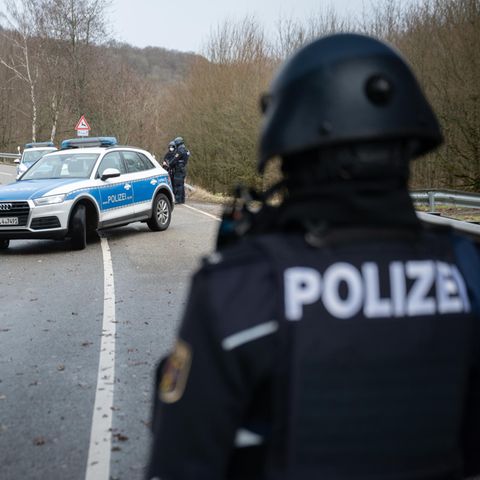 Ein Polizist in Schutzkleidung und mit Helmschaut auf ein diagonal über eine geschwungene Landstraße stehendes Polizeiauto