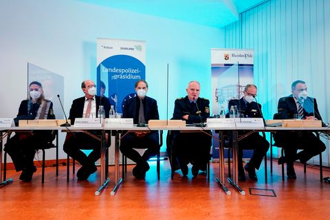 An zweier-Tischen sitzen links eine Frau und rechts von ihr fünf Männer in Anzügen oder Poluieuniformen