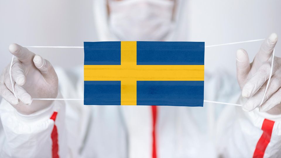 Ein Mann in Schutzanzug hält einen Mund-Nase-Schutz mit einer schwedischen Flagge nach vorne