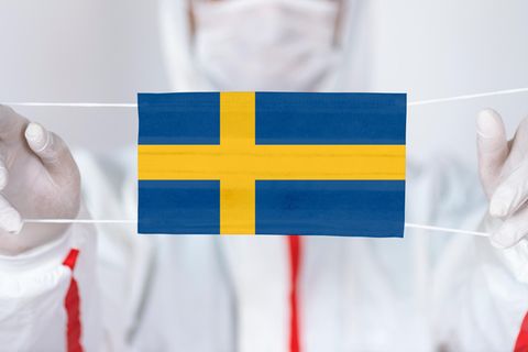 Ein Mann in Schutzanzug hält einen Mund-Nase-Schutz mit einer schwedischen Flagge nach vorne
