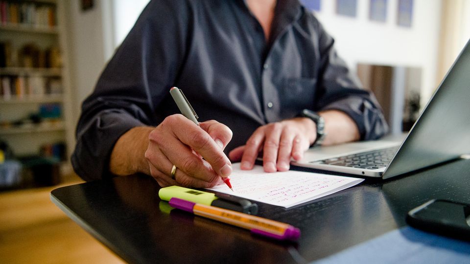 Ein Mann sitzt im Homeoffice an einem Schreibtisch und schreibt mit einem Stift etwas auf