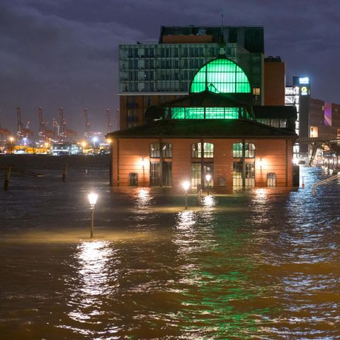 Ein Backsteingebäude mit grün beleuchteten Fenstern sowie altmodische Laternen ragen bei Dunkelheit aus braunem Wasser