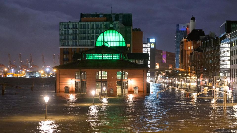 Ein Backsteingebäude mit grün beleuchteten Fenstern sowie altmodische Laternen ragen bei Dunkelheit aus braunem Wasser