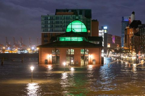 Ein Backsteingebäude mit grün beleuchteten Fenstern sowie altmodische Laternen ragen bei Dunkelheit aus braunem Wasser