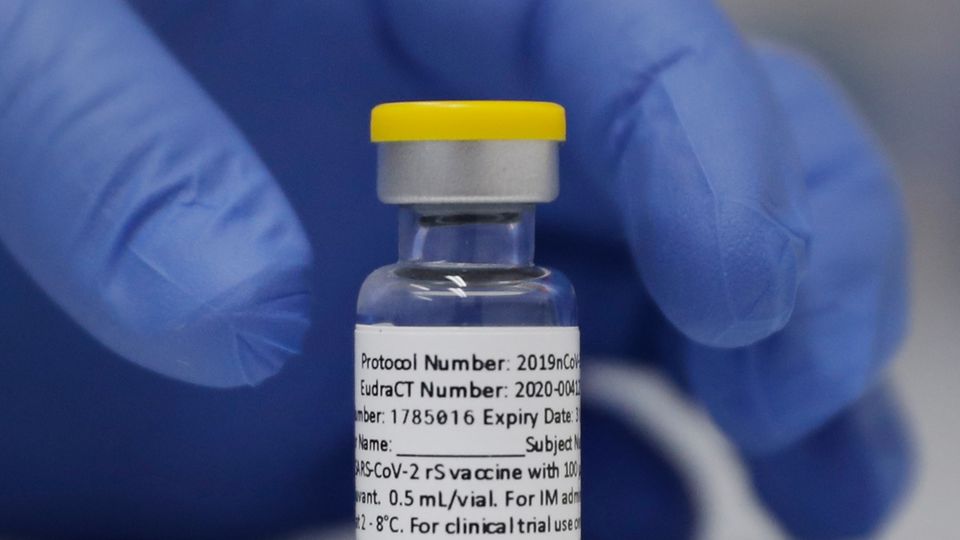 Eine linke Hand in blauem Gummihandschuh greift nach einer Ampulle mit Corona-Impfstoff von Novavax