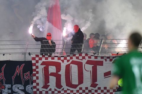 Schwarz gekleidete Männer mit roten Sturmhauben schwenken weiß brennende Pyrotechnik über einem Stadionzaun