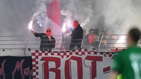 Schwarz gekleidete Männer mit roten Sturmhauben schwenken weiß brennende Pyrotechnik über einem Stadionzaun