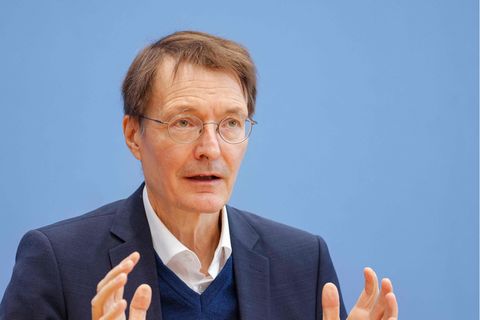 Ein weißer Mann mit braunem Seitenscheitel und runder Metallbrille gestikuliert vor einer blauen Wand