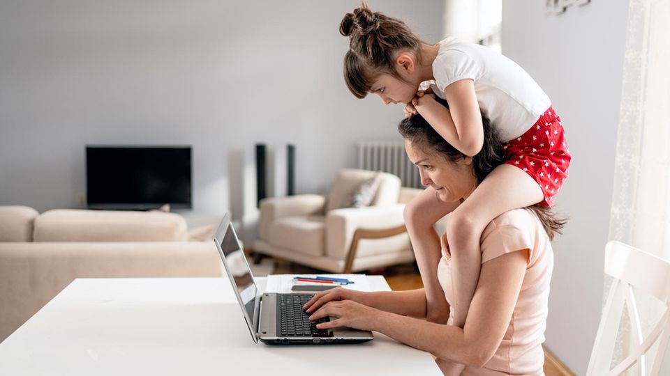 Mutter mit Kind auf den Schultern sitzt vor dem Computer