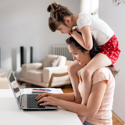 Mutter mit Kind auf den Schultern sitzt vor dem Computer