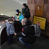 Neben einer Rolltreppe ist ein Mann mit Halbglatz in die Hocke gegangen, um mit einem Jungen auf Augenhöhe zu sprechen.