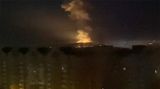 Auf einem pixeligen Foto ist Feuerschein und Rauch hinter einem langen Wohnblock zu erkennen