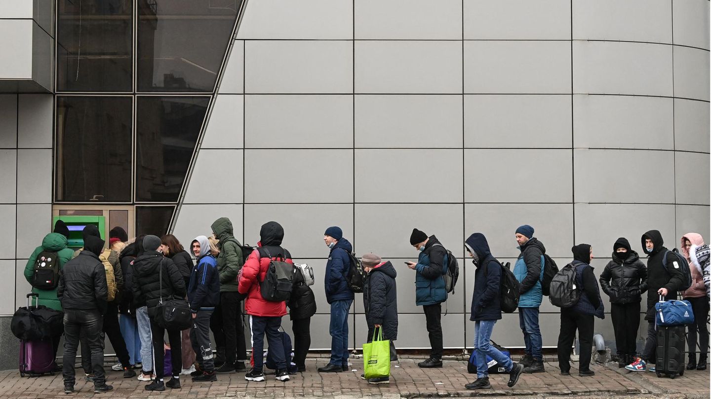 Vor einem grünen Geldautomaten stehen Menschen in Winterkleidung Schlange