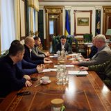 An einem langen, dunklen Holztisch sitzen sieben Männer, am Kopfende ein Mann in Anzug in grün gepolstertem Sessel