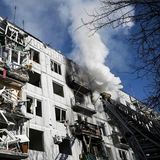Eine Drehleiter ragt an einem hellgrau verkleideten mehrstöckigen Haus mit zerstörten Balkonen empor. Weißer Rauch steigt auf