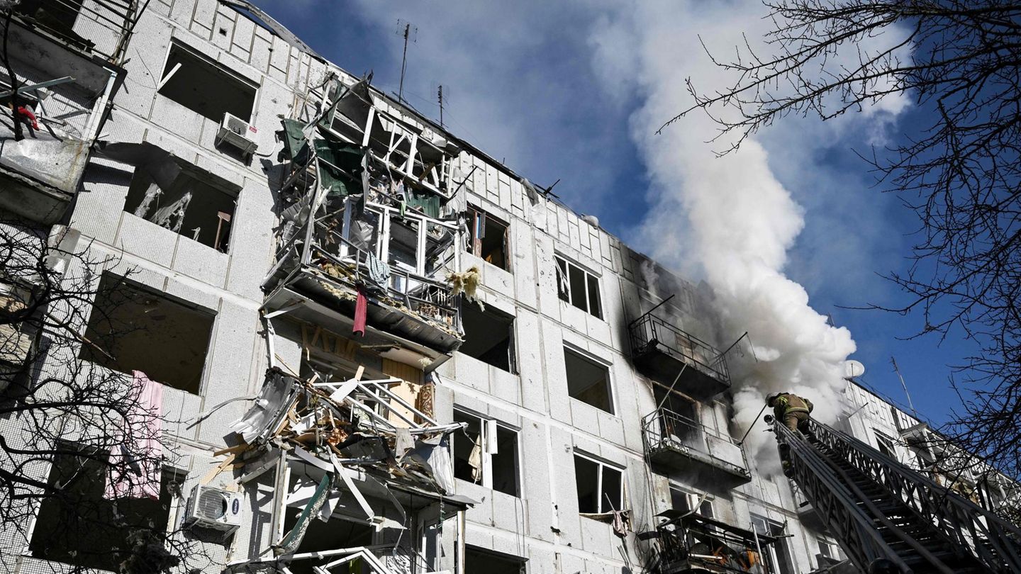 Eine Drehleiter ragt an einem hellgrau verkleideten mehrstöckigen Haus mit zerstörten Balkonen empor. Weißer Rauch steigt auf