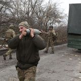 Ein Soldat trägt auf der linken Schulter eine graue Granate. Im Hintergrund steht ein Militär-Lkw