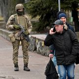 Soldaten in voller Kampfausrüstung stehen auf einer Straße, während zwei Männer in Winterkleidung von ihnen weggehen