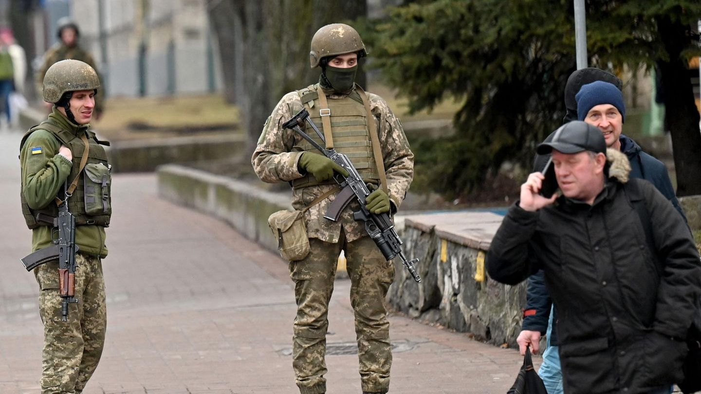 Soldaten in voller Kampfausrüstung stehen auf einer Straße, während zwei Männer in Winterkleidung von ihnen weggehen