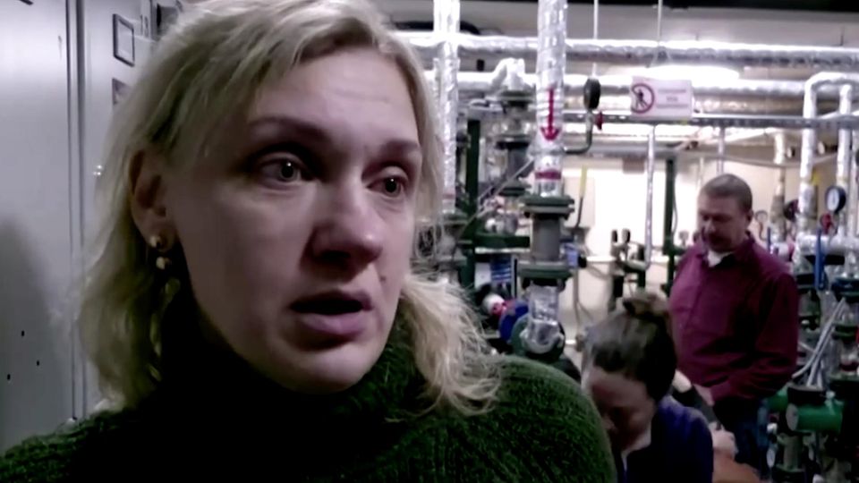 Eine müde wirkende weiße Frau mit schulterlangen blonden Haaren und dunkelgrünem Rollkragen-Pullover lehnt an einem Spind