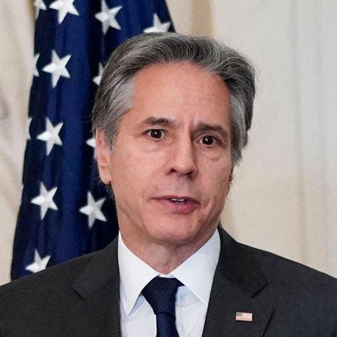 Ein weißer Mann mit grauem Seitenscheitel steht im dunklen Anzug und Krawatte vor drei unterschiedlichen Flaggen