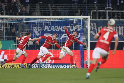 Vier Fußballspieler in roten Trikots und weißen Hosen laufen jubelnd über einen Fußballplatz