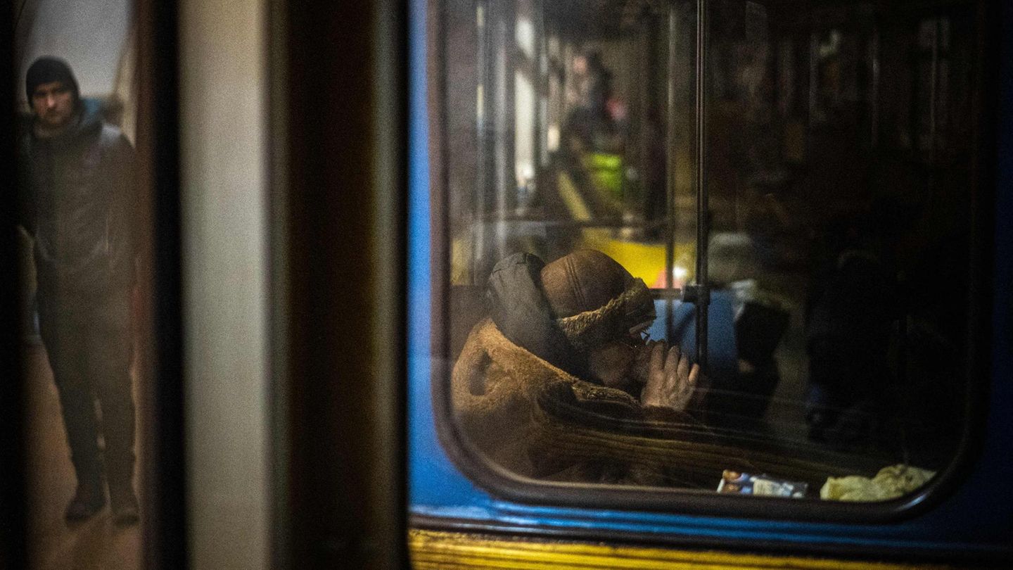 Ukraine Gesichter Krieg Frau Tee U-Bahn Hinter einem schmutzigen Fenster einer blau-gelb lackierten U-Bahn sitzt eine alte Frau im Wintermantel und trinkt Tee