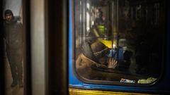 Hinter einem schmutzigen Fenster einer blau-gelb lackierten U-Bahn sitzt eine alte Frau im Wintermantel und trinkt Tee