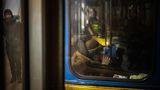 Hinter einem schmutzigen Fenster einer blau-gelb lackierten U-Bahn sitzt eine alte Frau im Wintermantel und trinkt Tee
