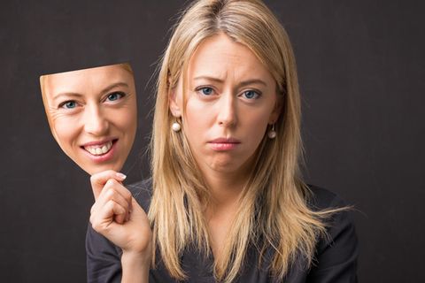 Eine Frau schaut traurig, in ihrer Hand hat sie eine Maske ihres lächelnden Gesichts
