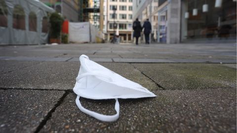 In einer Fußgängerzone in München liegt eine FFP2-Maske am Boden. Im Hintergrund gehe unscharf zwei Passanten weg