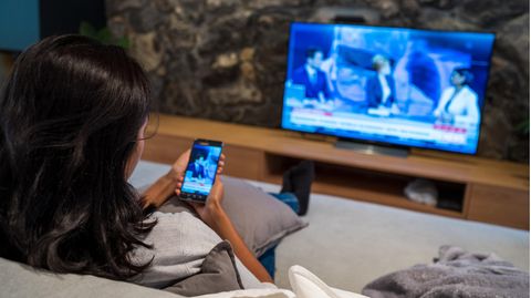 iPhone mit TV verbinden: Ein Frau sitzt auf dem Sofa und spiegelt das Display ihres iPhones auf dem TV