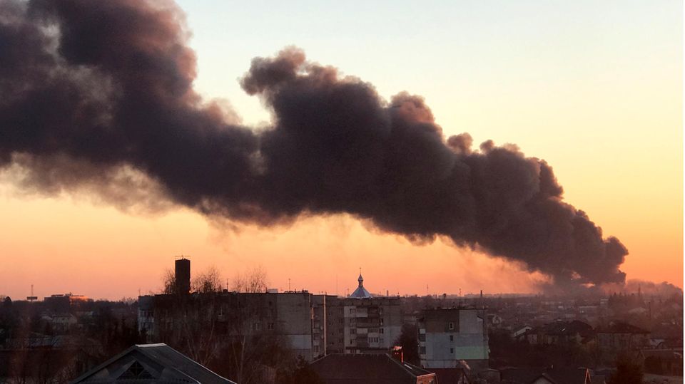 In der Morgendämmerung steigt über einer Stadt eine schwarze Rauchsäule von rechts unten nach links oben in den Himmel