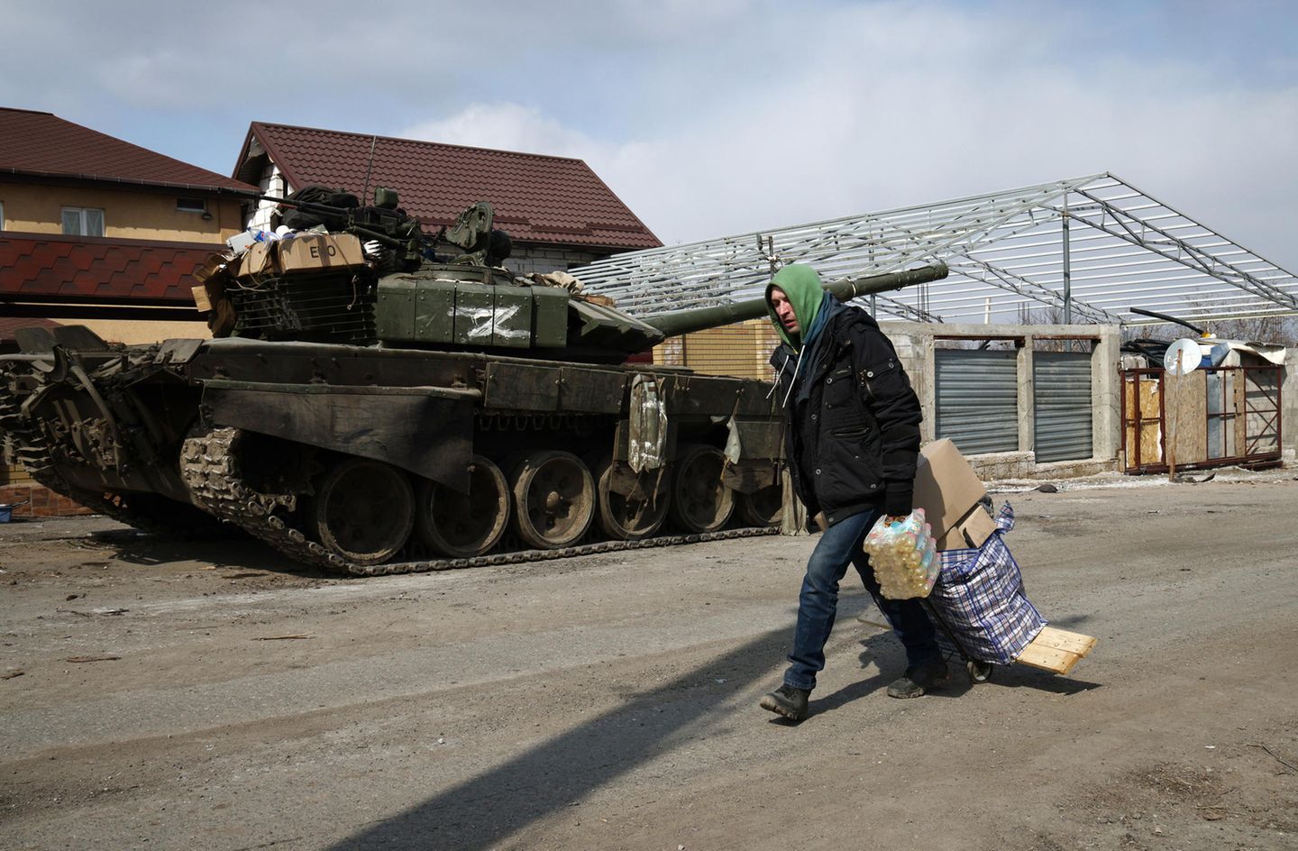 Ein Mann mit Kapuze trägt auf einer Straße Lebensmittel an einem Panzer vorbei