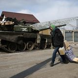 Ein Mann mit Kapuze trägt auf einer Straße Lebensmittel an einem Panzer vorbei