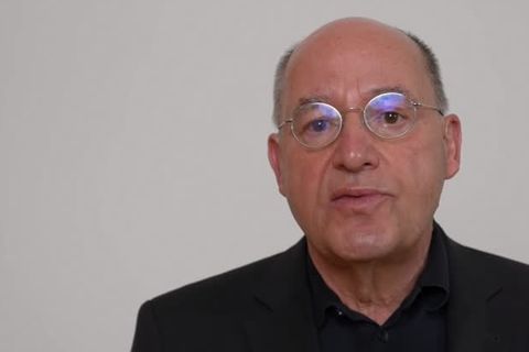 Ein weißer älterer Mann mit Halbglatze und runder Metallbrille sitzt in schwarzem Hemd vor hellgrauem Hintergrund