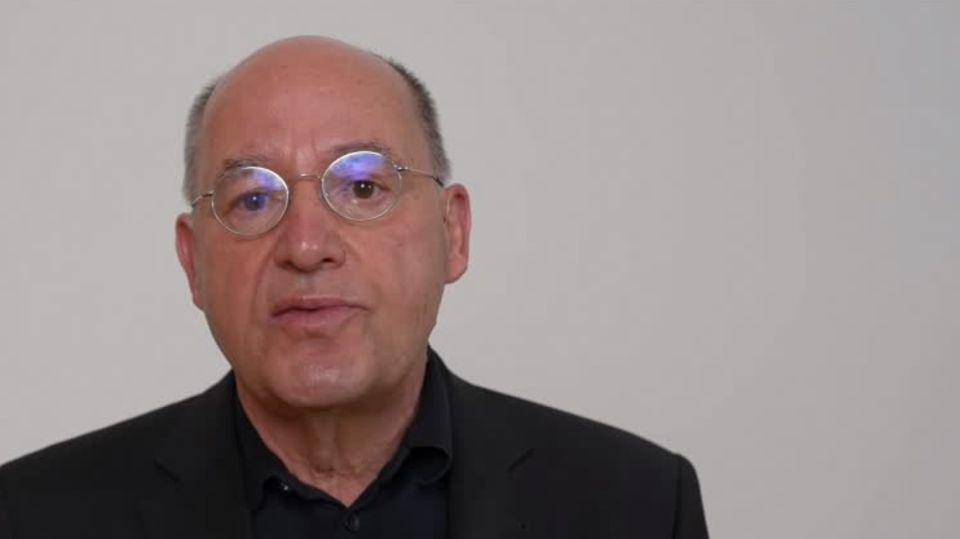 Gregor Gysi Ein weißer älterer Mann mit Halbglatze und runder Metallbrille sitzt in schwarzem Hemd vor hellgrauem Hintergrund