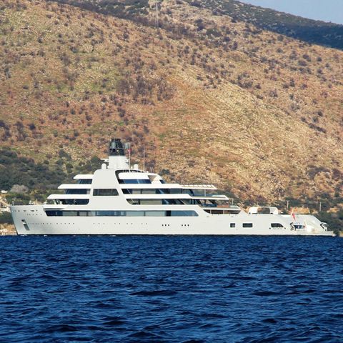 Eine riesige weiße Motoryacht liegt vor einer karg bewachsenen Küste in der Sonne