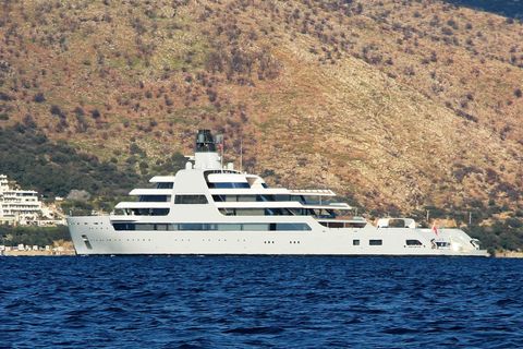 Eine riesige weiße Motoryacht liegt vor einer karg bewachsenen Küste in der Sonne
