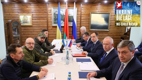 An einem langen Tisch sitzen links vier Männer in Uniformen, rechts fünf in Anzügen vor Flaggen der Ukraine und Russlands