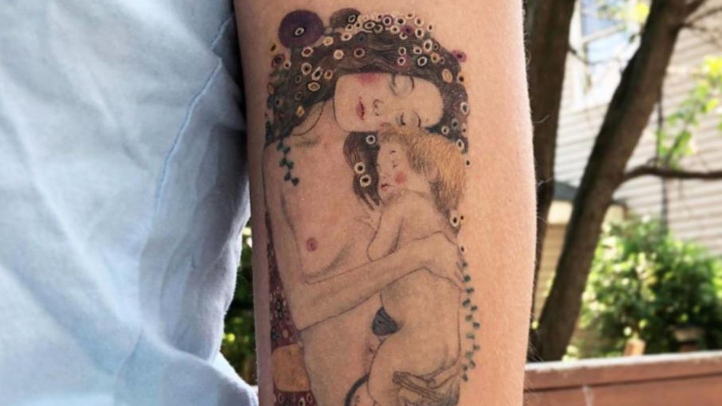 Eine nackte Mutter mit Blumen im Haar, die ihr schlafendes Kind eng umschlungen hält ist auf einen Arm tätowiert