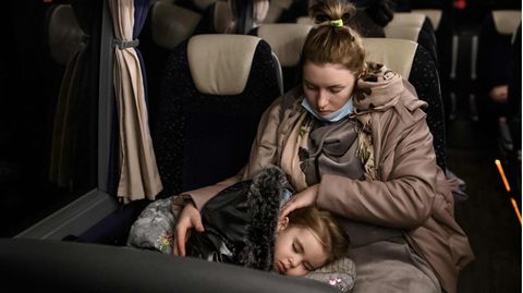 Eine weiße Frau in brauner Winterjacke sitzt in einem Bus und streichelt den Kopf eines Mädchens, das auf ihrem Schoß schläft