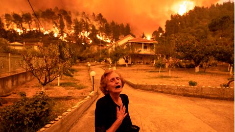 Eine hagere, alte Frau in Schwarz greift sich mit rechts ans Herz, während im Hintergrund Nadelwald in Flammen steht