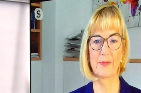 Links steht eine weiße junge Frau mit langen Haaren in einem TV-Studio, rechts auf einem Bildschirm spricht eine blonde Frau