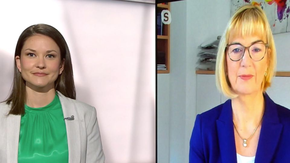 Links steht eine weiße junge Frau mit langen Haaren in einem TV-Studio, rechts auf einem Bildschirm spricht eine blonde Frau Links steht eine weiße junge Frau mit langen Haaren in einem TV-Studio, rechts auf einem Bildschirm spricht eine blonde Frau