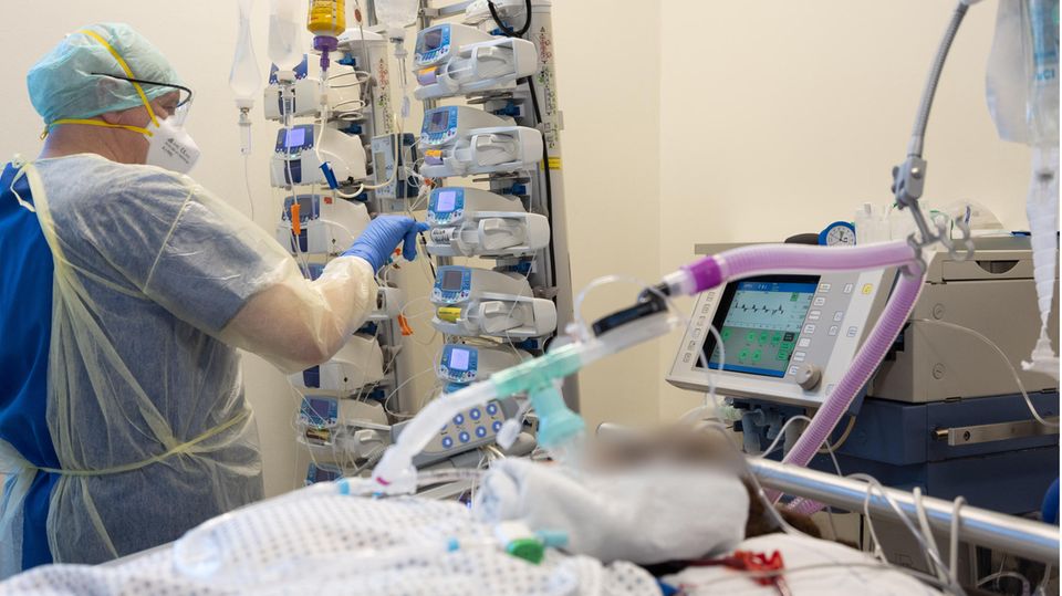 Ein weißer Mann in helblauer medizinischer Schutzkleidung drückt neben dem Bett eines beatmeten Patienten auf ein Gerät