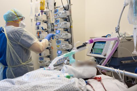 Ein weißer Mann in helblauer medizinischer Schutzkleidung drückt neben dem Bett eines beatmeten Patienten auf ein Gerät